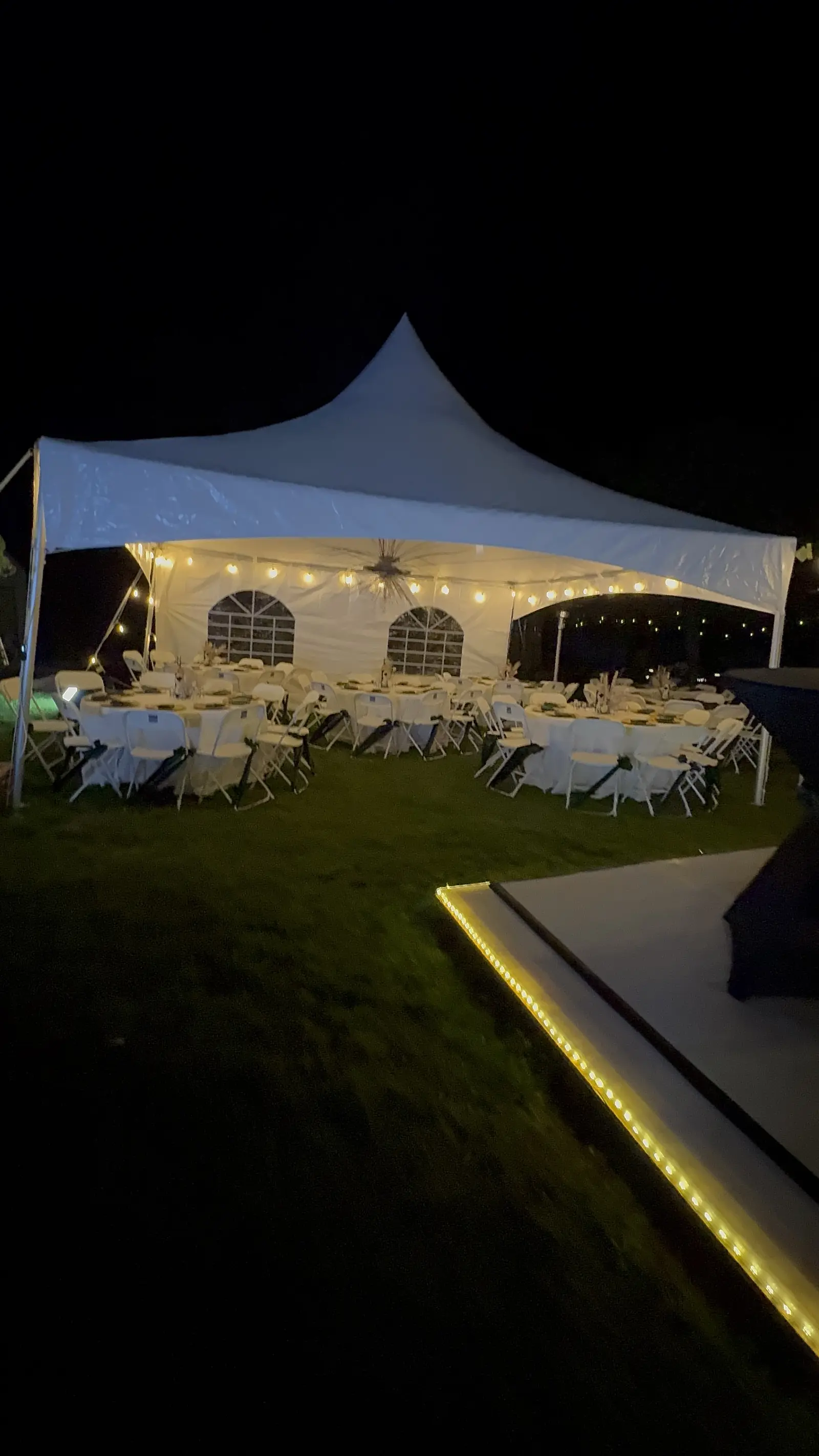 White high-peak tent lit with string lights at night, with set tables inside