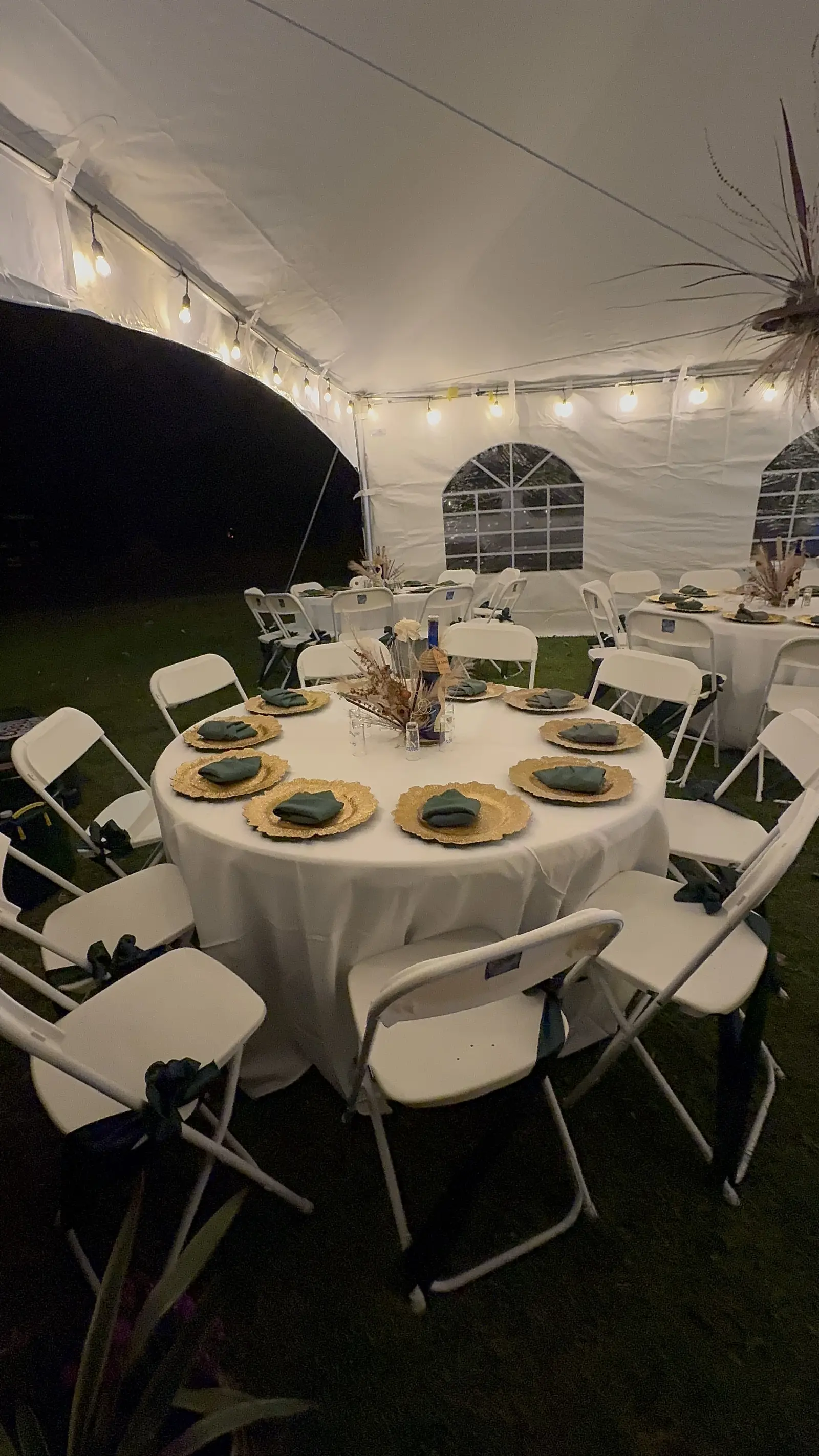 Round tables with white folding chairs and gold chargers inside a white tent lit with string lights